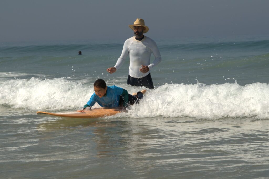 A kid finishing a wave lying on his belly during his first surf lesson, with surf instructor Ivo watching in the background.